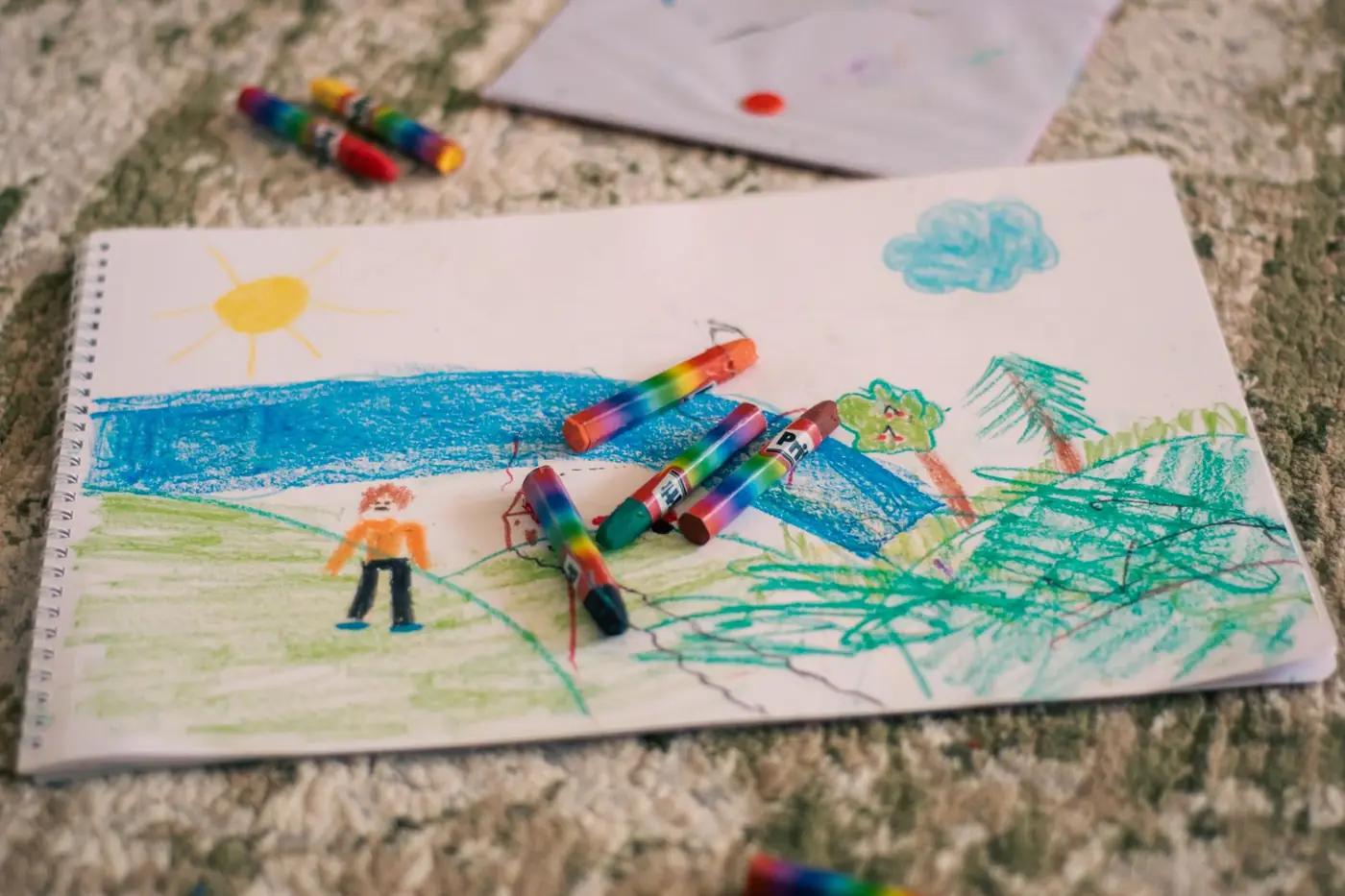 Il disegno a pastelli di un bambino, con il sole, il mare e gli alberi, e i pastelli appoggiati sopra